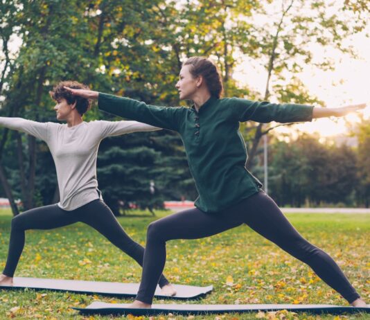 “벽을 넘어 숲으로”… 마음의 병 치유하는 ‘그린 엑서사이즈’ 열풍 Two women practicing yoga warrior pose outdoors.